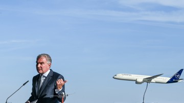 Lufthansa-Chef Carsten Spohr spricht bei der Taufe des Lufthansa Dreamliners vor einem fliegenden Flugzeug.