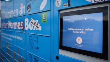 Nahaufnahme eines blauen Hermes-Paketautomaten mit einem Display auf dem steht: Willkommen an deiner Hermes Box.