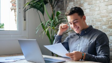 Ein junger Mann sitzt vor seinem Laptop und freut sich über das, was auf dem Papier in seiner Hand steht - er hat freudig eine Faust erhoben