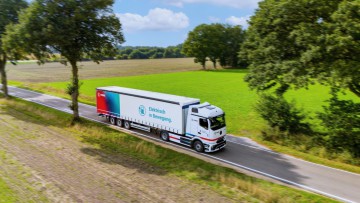 Der eActros 600 von Mercedes Benz im Branding des Logistkdienstleisters Fiege fährt auf einer Straße durch eine Parklandschaft mit grünen und braunen Äckern sowie Bäumen und im Hintergrund ein Wald. Auf dem Trailer steht neben dem Fiege-Logo auf einem Far