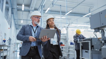 Zwei professionelle Ingenieure der Schwerindustrie, die in der Fabrik Schutzhelme tragen. Gehen und diskutieren Industriemaschinenanlagen, Arbeiten am Laptop.