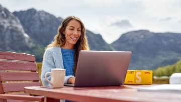 Frau an einem Tisch in der Natur lächelt in ihren Laptop, neben ihr ein DHL Paket