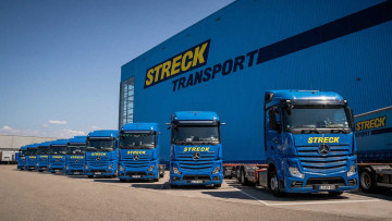 Neun Lkw des Fuhrparks stehen aufgereiht am Standort Freiburg von Streck Transport vor einer großen Halle. Die Lkw sind blau genau wie die Halle. Oben links auf der Halle steht der Name des Unternehmens in gelber Schrift, auf den Lkw steht er vorne 