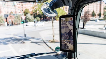 Im Bild der Blick nach rechts aus dem Innenraum des Fahrerhauses im Lkw mit Fokus auf einen Abbiegeassistenten von Mercedes-Benz, der grade einen E-Scooter Fahrer neben dem Lkw anzeigt und über ein kleines rotes Warndreieck auf die Gefahr aufmerksam macht