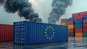 Symbolbild für den CO2-Grenzausgleichsmechanismus der EU: Ein großer blauer Frachtcontainer mit der EU-Flagge auf der Seite steht auf einem Lagerplatz in einem Containerhafen – weitere verschiedenfarbige Container sind im Hintergrund zu sehen. Aus dem bla