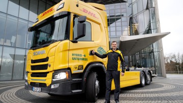 Tobias Meyer, CEO DHL Group, steht vor dem Testfahrzeug, einer Scania-Sattelzugmaschine in gelbem Design mit DHL-Logo und grünem Schriftzug „Hybrid unterwegs“