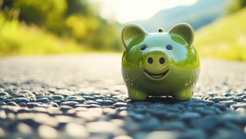 Ein grünes Sparschwein steht auf einer Landstraße, im Hintergrund ist verschwommen eine grüne Bergkette zu erkennen. Symbolbild für Finanzierung nachhaltiger Logistik- und Transportlösungen und entsprechende Fördergelder