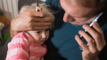 Ein Vater hält seine kranke Tochter im Arm, in der einen Hand ein Fieberthermometer, mit der anderen hält er sein Mobiltelefon ans Ohr