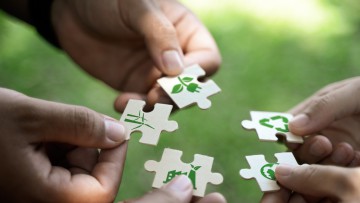Hände verschiedener Personen halten Puzzleteile in einem Kreis, auf denen Symbole für eine CO2-arme oder -neutrale Wirtschaft abgebildet sind, darunter ein Windrad, ein Stecker mit grünen Blättern und das Symbol für Kreislaufwirtschaft