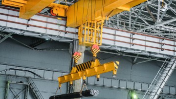 Ein gelber Brückenkran transportiert Metallstäbe als Fracht in einer Halle. 