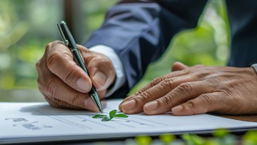 Ein Geschäftsmann unterschreibt ein Dokument, auf dem ein grünes Blättchen einer Pflanze liegt, im Hintergrund sieht man die Bäume eines Waldes durchs Fenster scheinen