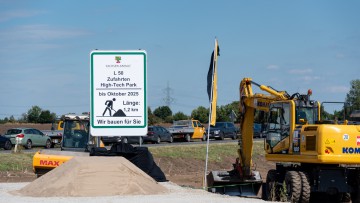 Baumaschinen stehen an der Zufahrt zu einem Acker. Links daneben weist ein Schild auf den Ausbau der Landesstraße 50 hin. Von dieser Landesstraße werden Zufahrten zum High-Tech-Park auf dem Eulenberg abzweigen. Dort wollte der US-Chiphersteller zwei Halbl