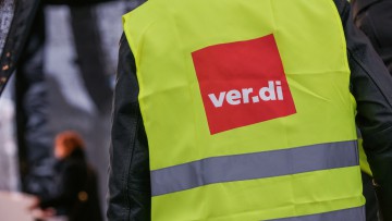 Detailaufnahme: Ein Beschäftigter mit einer Warnweste, auf der ein Verdi-Logo abgebildet ist, steht im Vordergrund (Symbolfoto)