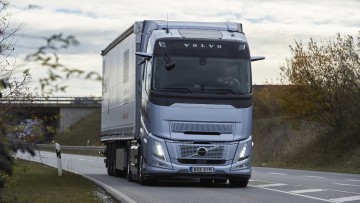 Ein Volvo FH Aero I-Save fährt bei bedecktem Himmel und schummerigen Lichtverhältnissen auf einer Straße durch die Landschaft, von vorne aufgenommen, zu sehen ist eine buschbestandene Böschung und im Hintergrund eine Brücke