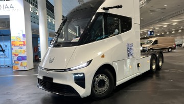 Der E-Lkw fährt in einer Lagerhalle. Die weiß lackierte Dreiachs-Sattelzugmaschine ist ohne Auflieger unterwegs 