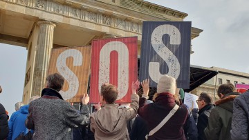 Drei Leute halten vor dem Brandenburger Tor Schilder hoch, die SOS ergeben