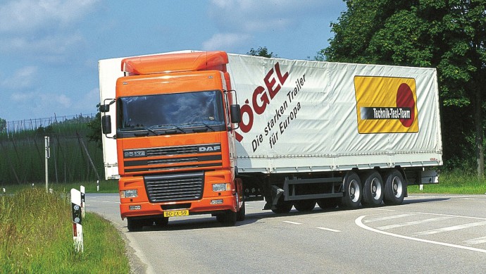 Ein in Orange lackierter DAF XF 95 beim Abbiegevorgang auf einer Landstraße. Das Fahrzeug hat einen Sattelauflieger aufgesattelt