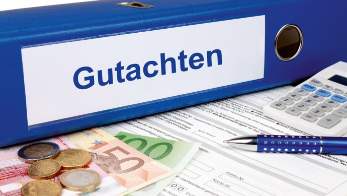 Ein blauer Ordner mit Aufschrift "Gutachten" liegt auf Papierstapel, daneben Taschenrechner, Geld, Kugelschreiber