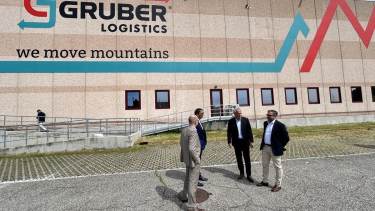 Vier Männer im Anzug stehen auf dem Asphalt vor einem großen braunen Gebäude mit der Aufschrift Gruber Logistics über dem Slogan we move mountains