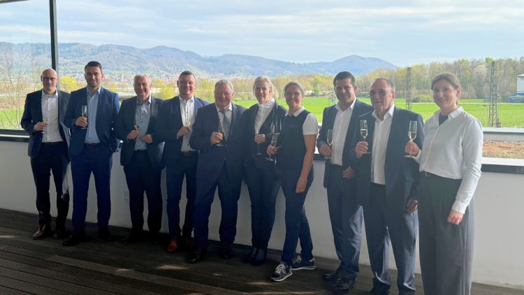 Männer und Frauen stehen in schicker Kleidung mit einem Glas Sekt in der Hand vor einem Fenster mit Ausblick auf eine sonnige Hügellandschaft