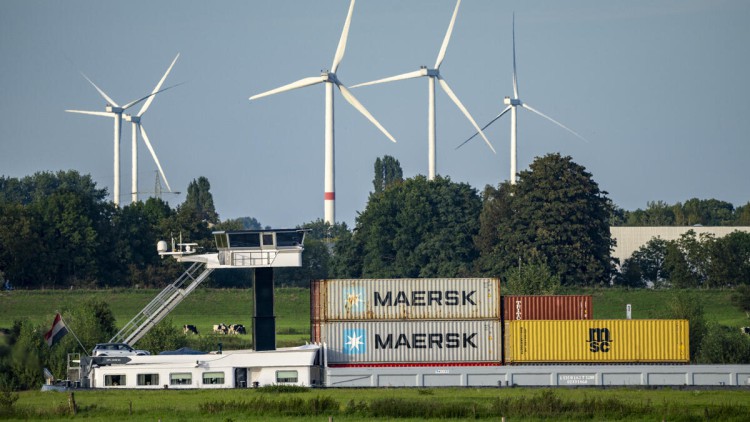 Beladenes Container Frachtschiff auf einem Fluss vor grünen Wiesen und einem Windpark