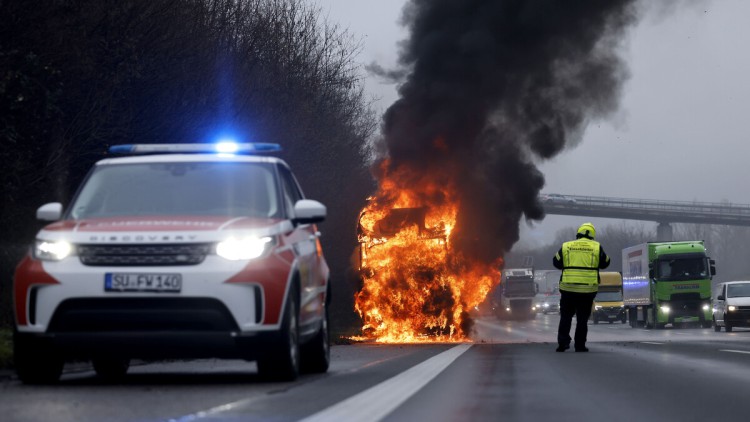 Ein brennender Lkw steht auf einer Autobahn, im Vordergrund Sicherheitskräfte