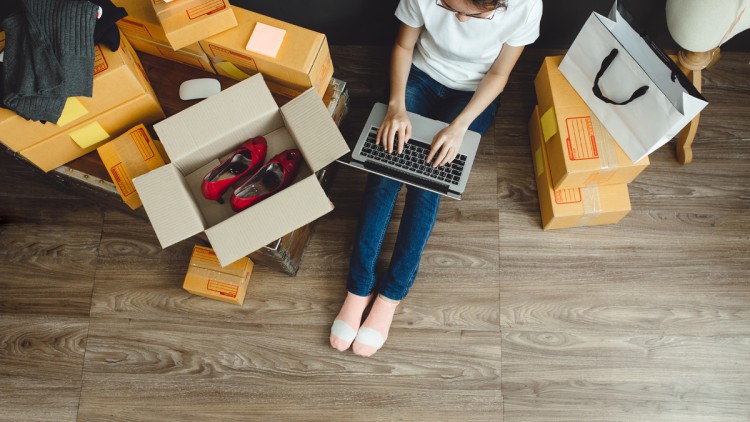 Frau sitzt auf dem Boden am Laptop zwischen gepackten und noch offenen Paketen
