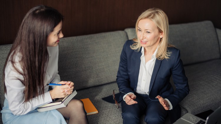 Aus der Vogelperspektive: Eine blonde Frau spricht und eine braunhaarige Frau hört ihr zu, beide sitzen auf einer grauen Couch