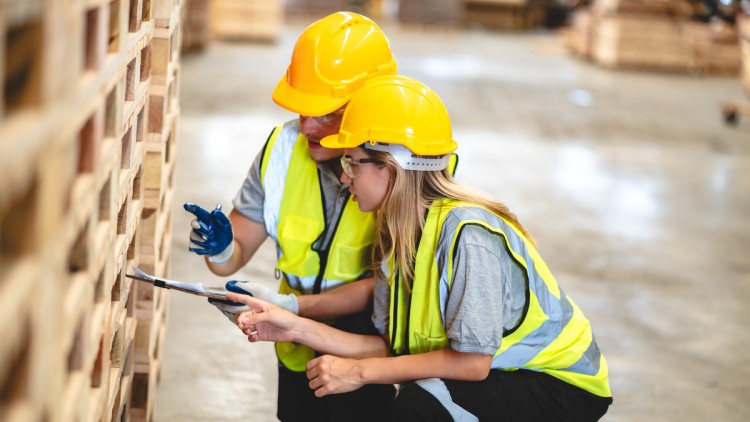 Ein Mann mit Warnweste und Klemmbrett sitzt in der Hocke neben einer jungen Frau in Warnweste und erklärt ihr etwas