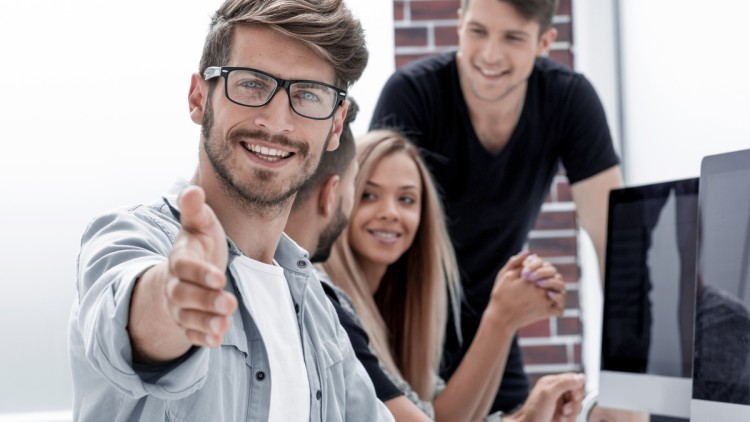 Bebrillter Mann im Büro streckt lächelnd die Hand in die Kamera, schwatzende Kollegen im Hintergrund