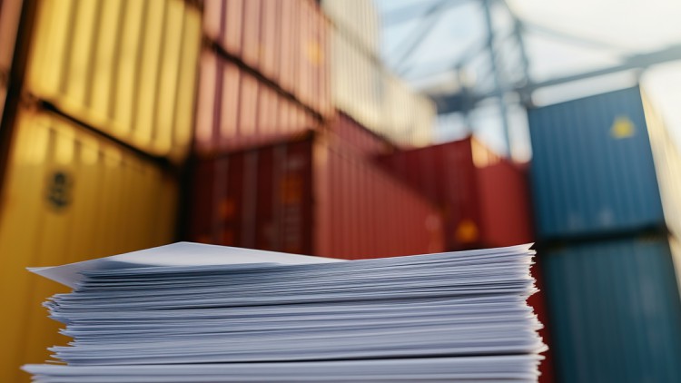 Ein Stapel Papiere liegt im Vordergrund. Im Hintergrund sind verschwommen verschiedene Container in unterschiedlichen Farben zu sehen