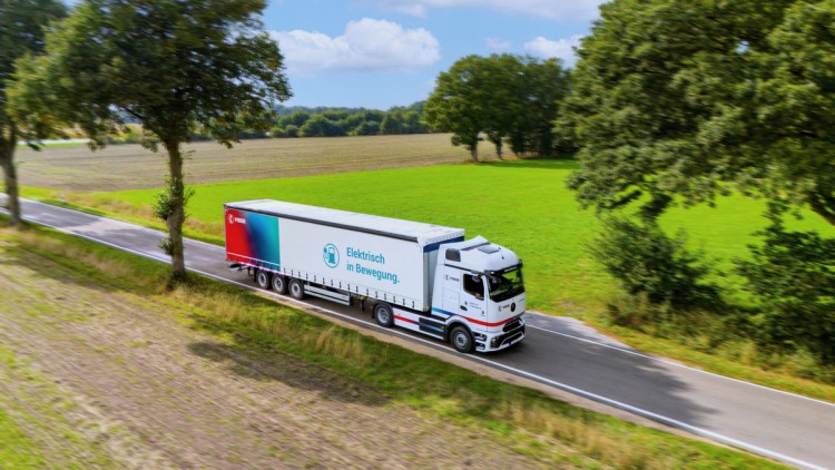 Der eActros 600 von Mercedes Benz im Branding des Logistkdienstleisters Fiege fährt auf einer Straße durch eine Parklandschaft mit grünen und braunen Äckern sowie Bäumen und im Hintergrund ein Wald. Auf dem Trailer steht neben dem Fiege-Logo auf einem Far