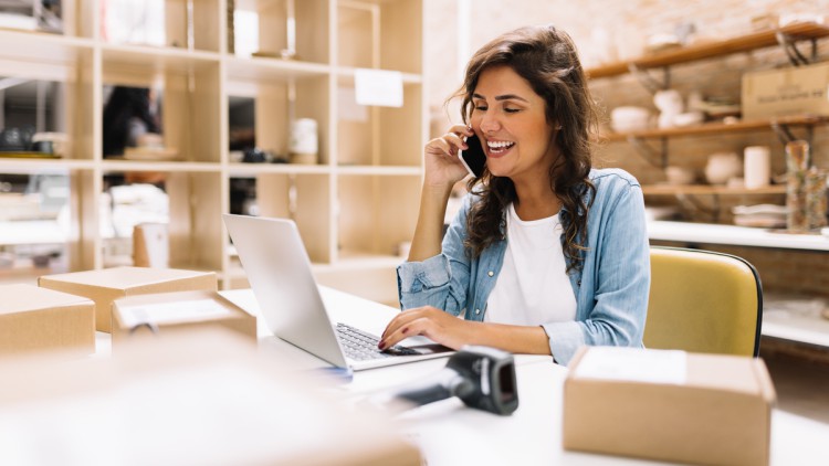 Lächelnde Unternehmerin telefoniert, während sie an einem Laptop in ihrem Lager arbeitet