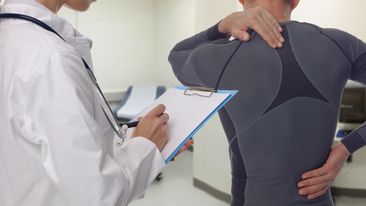 Männlicher Patienten fasst sich an den Rücken während der ärztlichen Untersuchung einer neben ihm stehenden Ärztin