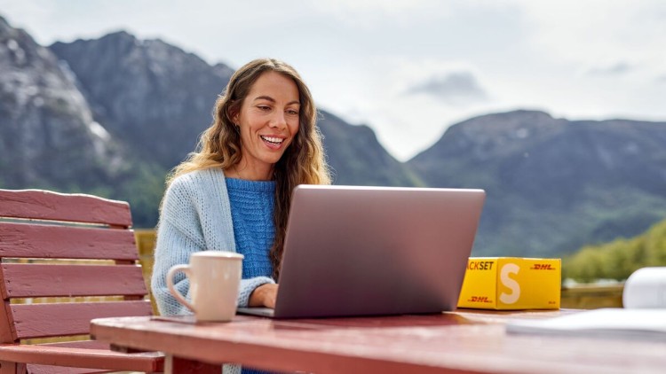 Frau an einem Tisch in der Natur lächelt in ihren Laptop, neben ihr ein DHL Paket