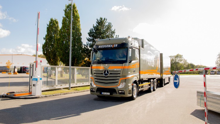 Ein Lkw der Niedermaier Spedition fährt bei strahlendem Sonnenschein von einem Betriebshof in einem Gewerbegebiet. Links vom Lkw ist eine Lagerhalle mit Entladerampen  und klein ein dort grade stehender Lkw zu sehen