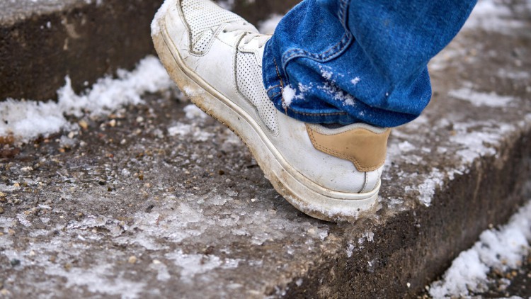 Ein Fuß einer Person tritt in den Schnee auf eine vereiste Treppe. Die rutschige Oberfläche und die dünne vereinzelt angetaute Schneeschicht symbolisieren die Gefahr von Glätte und Ausrutschen im Winter