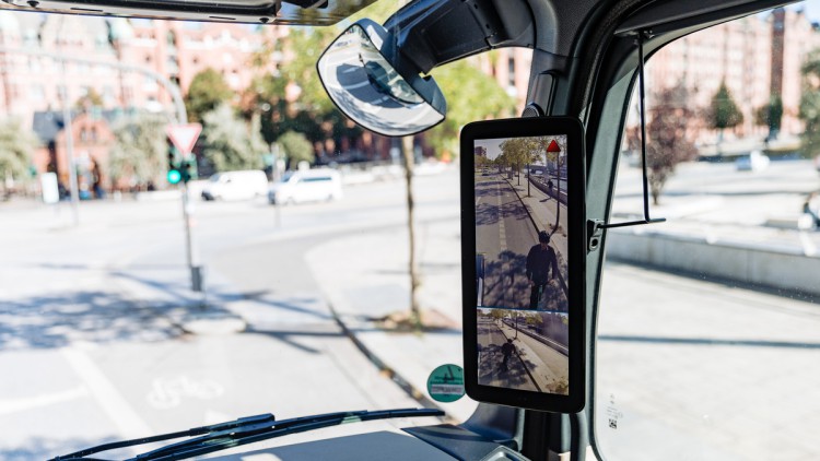 Im Bild der Blick nach rechts aus dem Innenraum des Fahrerhauses im Lkw mit Fokus auf einen Abbiegeassistenten von Mercedes-Benz, der grade einen E-Scooter Fahrer neben dem Lkw anzeigt und über ein kleines rotes Warndreieck auf die Gefahr aufmerksam macht