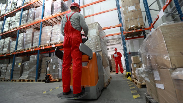 Ein Staplerfahrer mit roter Arbeitslatzhose fährt durch ein Lager mit Regalen und verpackten Kartons