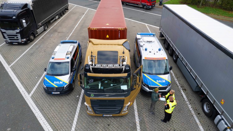 Kontrolle eines Lkw auf einem Rastplatz: Zwei Beamte des BALM stehen auf dem Parkplatz für Lkw und überprüfen die Papiere des Lkw-Fahrers.