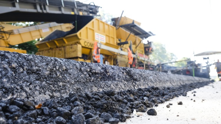 Asphaltreste liegen neben der Autobahn 215 zwischen dem Autobahndreieck Bordesholm (Schleswig-Holstein) und der Anschlussstelle Blumenthal. Im Hintergrund arbeiten ein Asphaltaufbringer und Arbeiter. Auf dem Teilstück wurde 2021 Kompaktasphalt aufgetragen