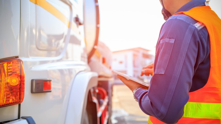 Mann in Warnweste steht neben Lkw und hält ein Tablet in der Hand, im Hintergrund ist ein Gebäude zu sehen