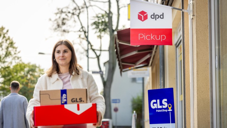 Eine Frau steht mit einem Paket von DPD und einem von GLS vor einem Paektshop, vor dem beide Marken als Fahnen beziehungsweise Aufsteller auf das Angebot des Shops für die Paketannahme und -abgabe hinweisen (Symbolbild)