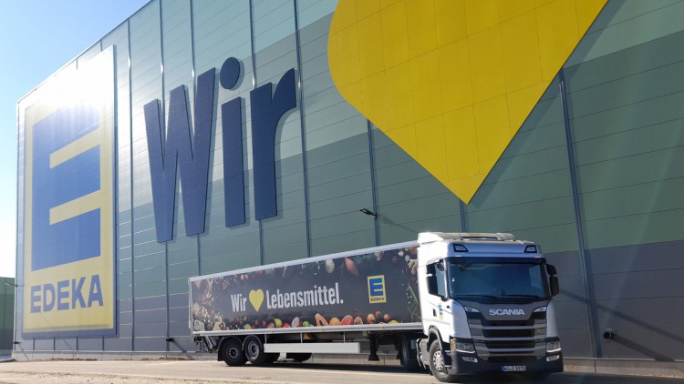 Ein Lkw von Edeka steht vor einem großen Markt mit riesiger Glasfront und großem Logo  sowie dem Slogan des Unternehmens mit dem gelben Herz.Die Umstellung der Flotte von Edeka Nordbayern-Sachsen-Thüringen auf CNG-Lkw beginnt am neuen Logistikzentrum am S