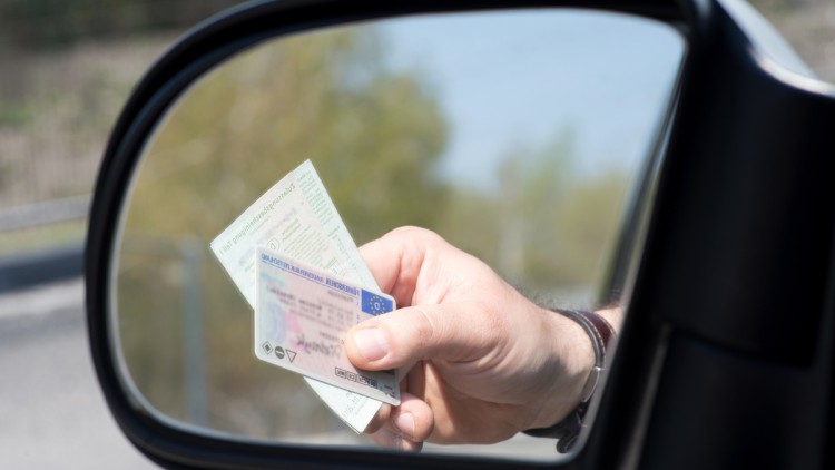 Im Bild sieht man in einem Rückspiegel eine Hand, die Führerschein und Fahrzeugpapiere aus dem Fenster hält, möglicherweise im Rahmen einer Kontrolle
