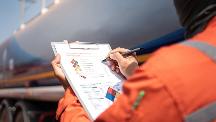 Eine Person in orangefarbener Warnschutzkleidung hält vor einem Tankauflieger ein Klemmbrett mit einer Checkliste für Gefahrstoffe. Auf dem Formular sind verschiedene Gefahrgutkennzeichen und Sicherheitsinformationen sichtbar.