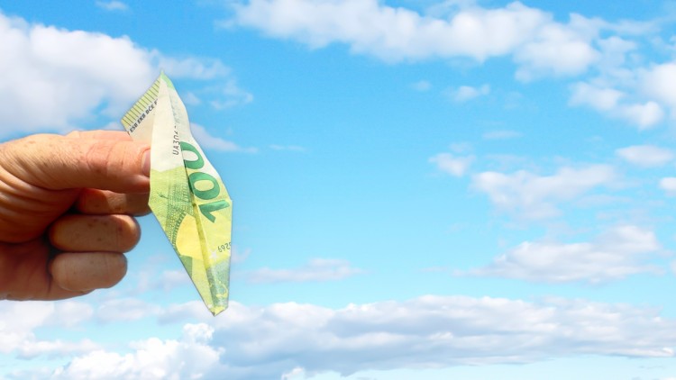 Eine Hand hält einen 100 Euro Schein in Form eines Papierfliegers nach unten, als würde dieser in den Sturzflug gehen, Hintergrund ist ein blauer Himmel mit Wolken