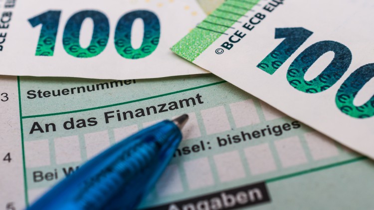 Ein Kugelschreiber liegt auf einem grünen Formular für Steuererklärungen, Großteils verdeckt von zwei 100-Euro-Scheinen. Zu lesen ist unter anderem: „An das Finanzamt“