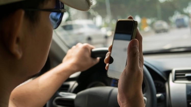 Symbolbild Handyverstoß: Im Bild sieht man die Nahaufnahme eines Mannes, der während der Fahrt mit einem Auto sein Smartphone benutzt