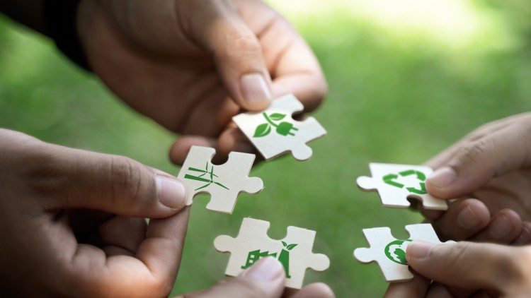 Hände verschiedener Personen halten Puzzleteile in einem Kreis, auf denen Symbole für eine CO2-arme oder -neutrale Wirtschaft abgebildet sind, darunter ein Windrad, ein Stecker mit grünen Blättern und das Symbol für Kreislaufwirtschaft
